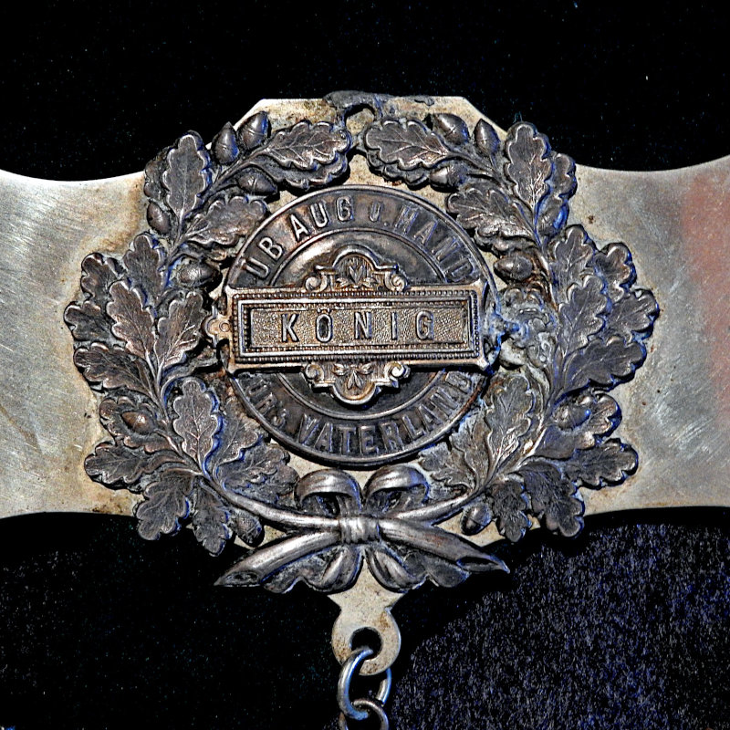  Das Kettenschild mit der Bezeichnung „KÖNIG“ und dem Motto: „ÜB AUG UND HAND FÜR'S VATERLAND“.